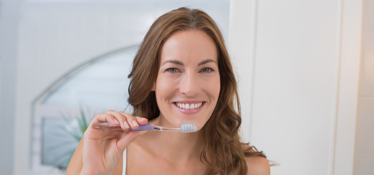 Eine Frau mit Zahnbürste in der Hand lächelt in die Kamera. 