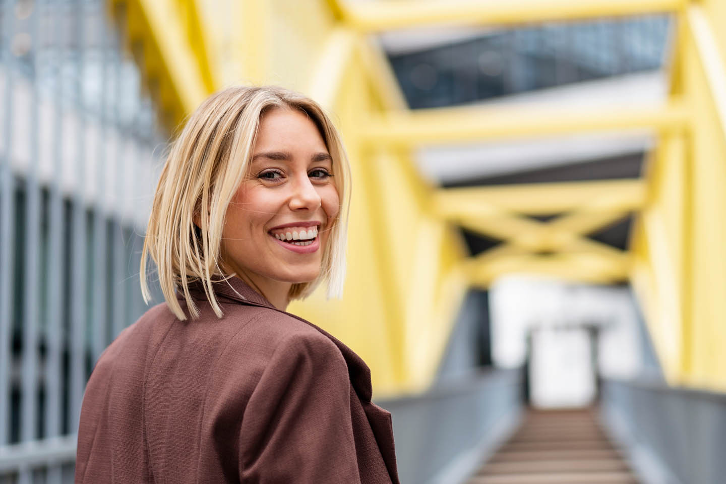 Eine Blondine mit Bob lächelt über die Schulter während sie über eine Brücke geht. 