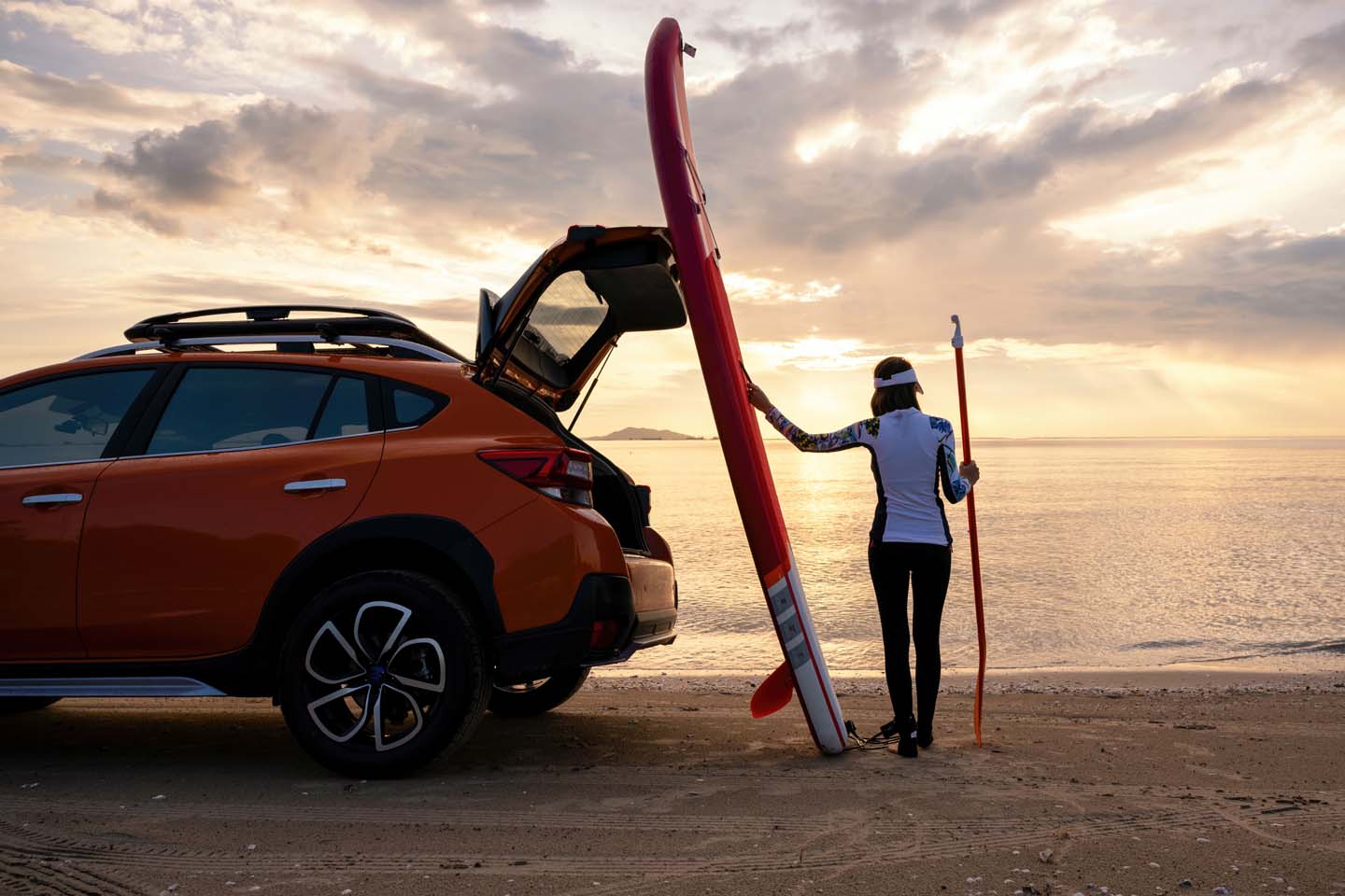 Ein Standup-Paddler steht mit seinem ans Auto gelehnten Board am Strand. Er betrachtet den Sonnenaufgang. 
