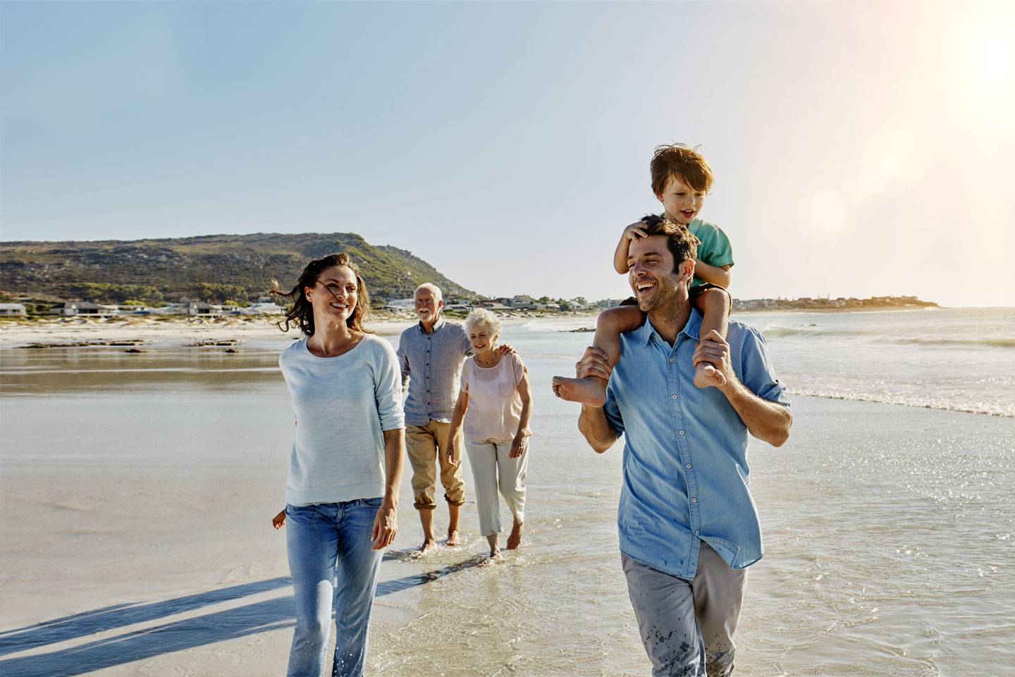 Drei Generationen – Großeltern, Eltern und Kind – gehen lachend am Strand spazieren.