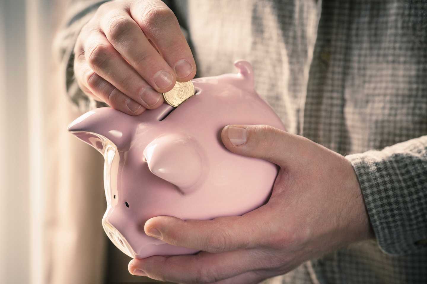 Ein Sparschwein wird von einem Mann in der einer Hand gehalten und die andere Hand steckt ein Geldstück hinein.