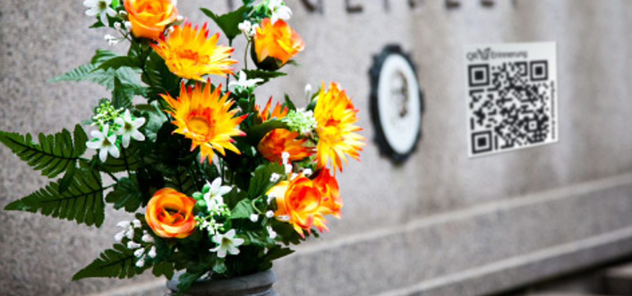 Ein Blumengesteckt steht vor einem Grabstein. Auf diesem befindet sich ein QR-Code.