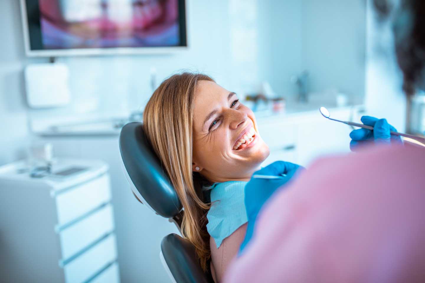 Frau lacht und sitzt auf einem Zahnarztstuhl. Ein Zahnarztspiegel wird vom Zahnarzt in der Hand gehalten.