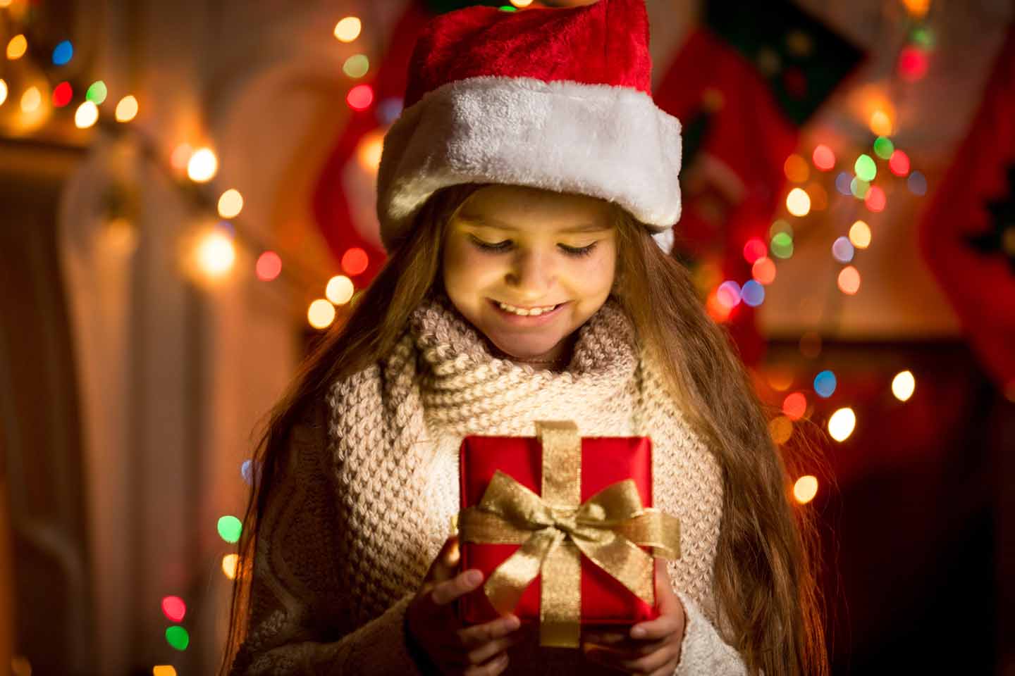 Ein kleines Mädchen mit Weihnachtsmütze öffnet ein Geschenk. 