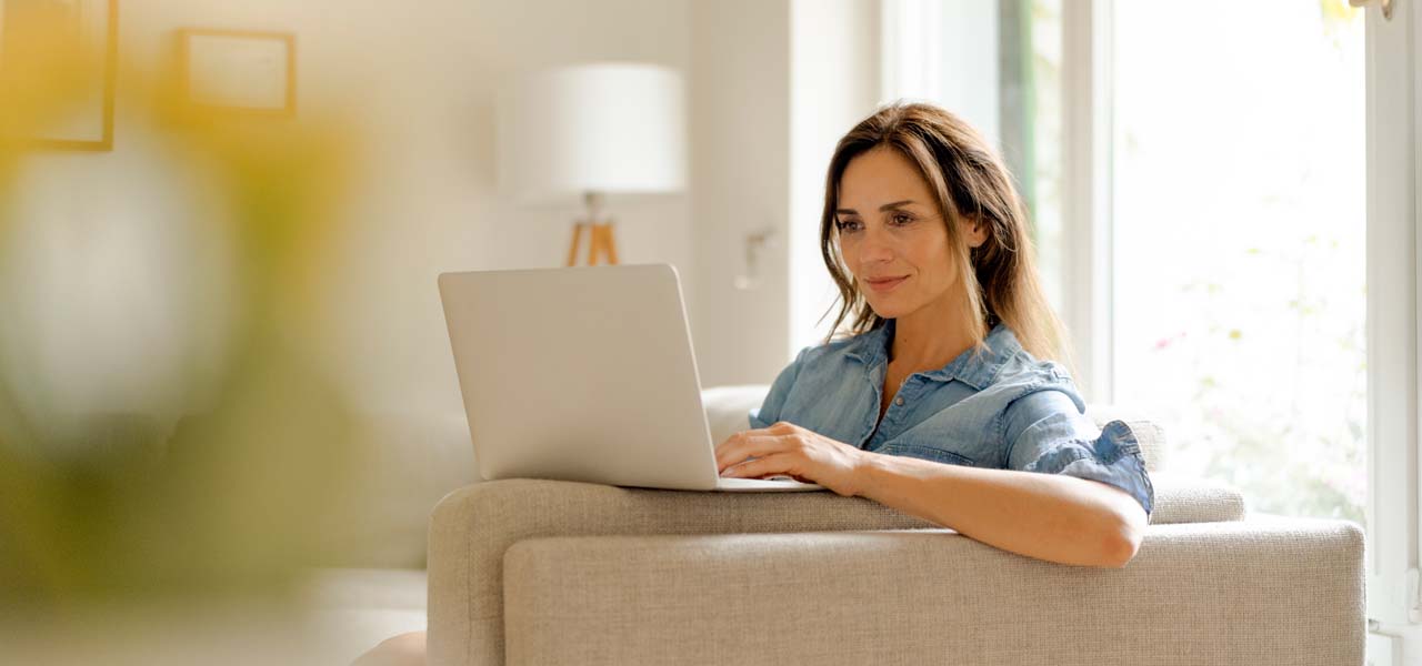 Eine Frau sitzt auf dem Sofa in einem gemütlichen Zuhause und schaut in ihren Laptop.