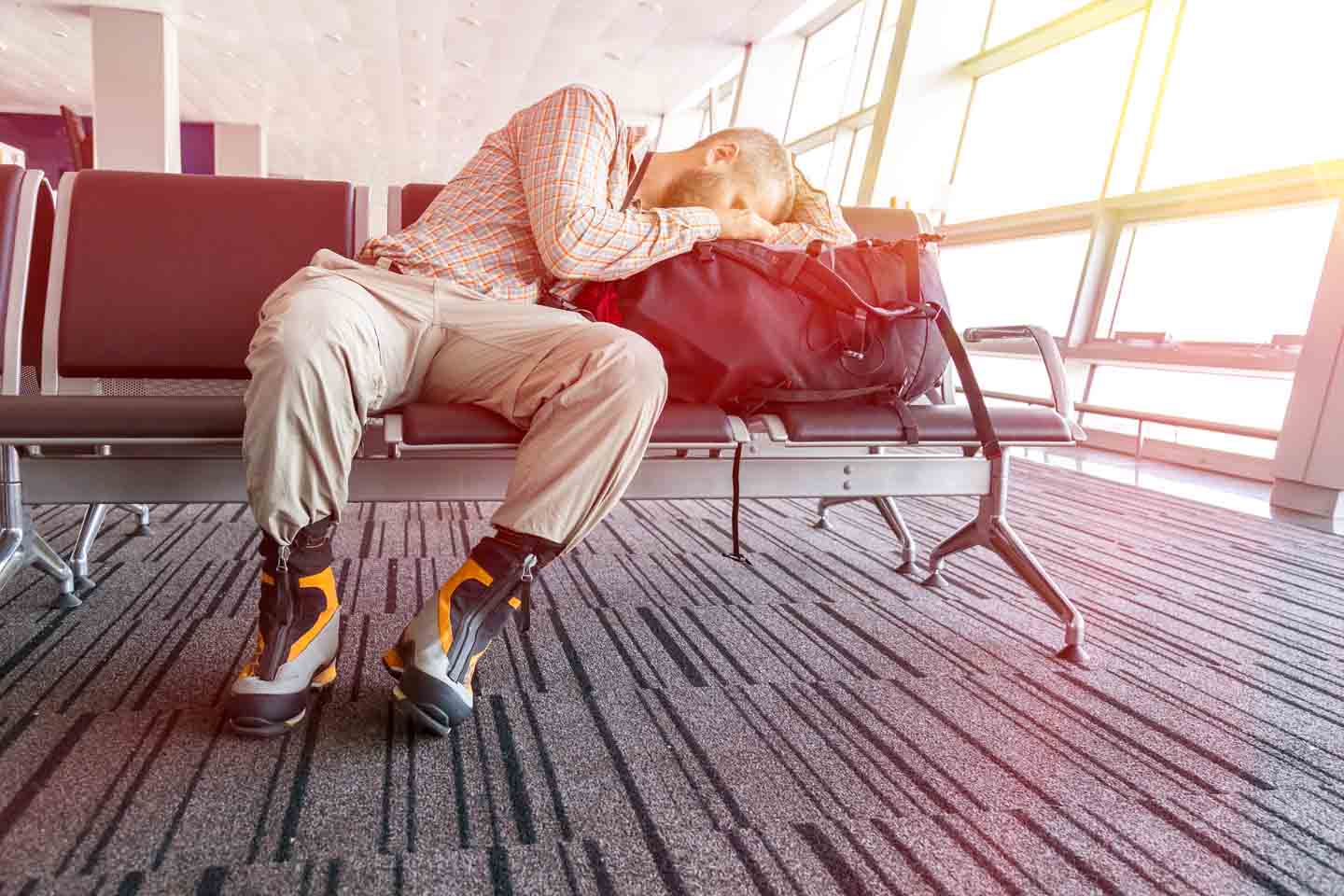 Ein Mann schläft halb sitzend, halb liegend auf einer Bank in der Wartehalle eines Flughafens.