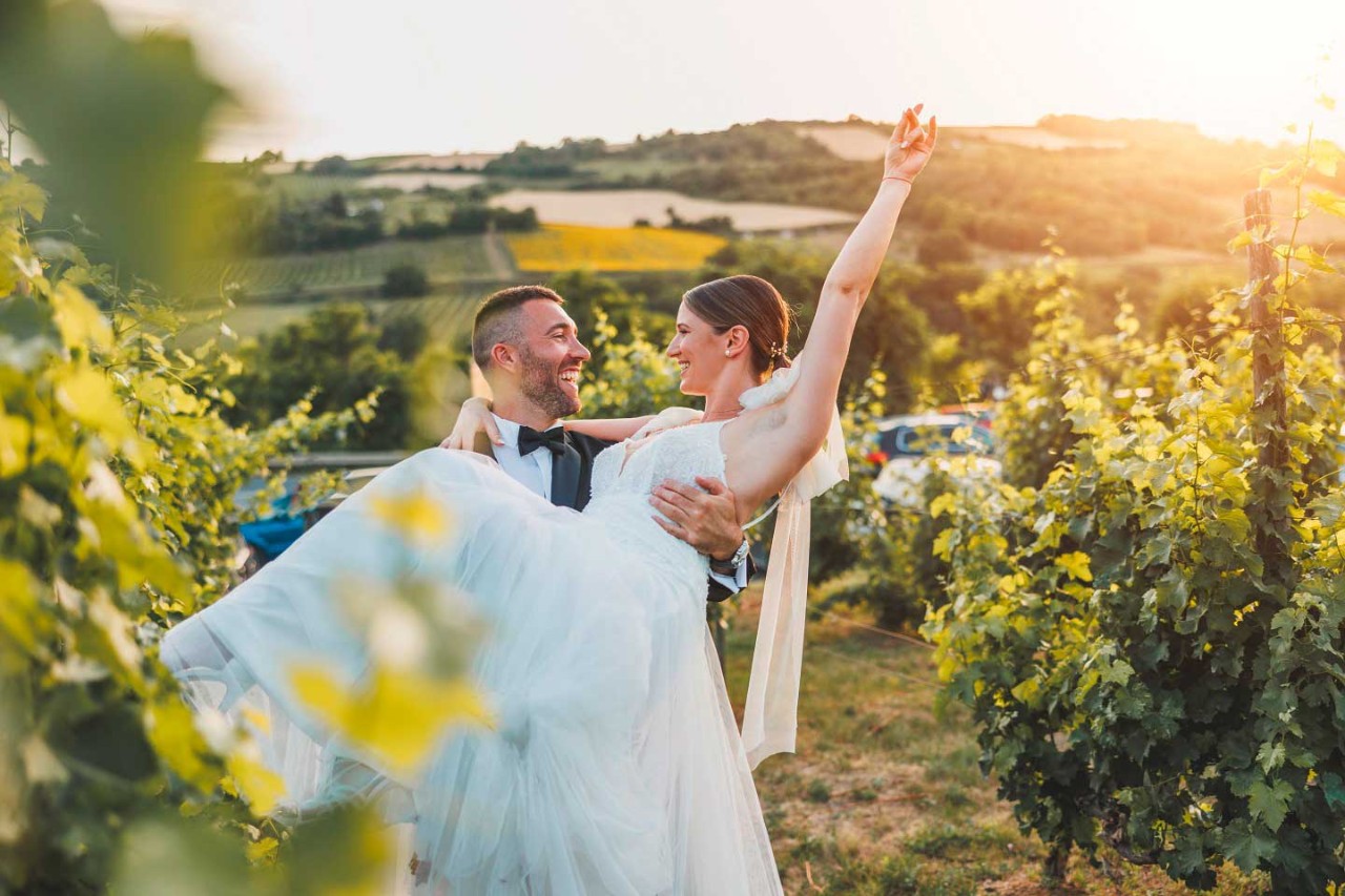 Ein Bräutigam trägt eine Frau im Hochzeitskleid durch einen Weinberg.