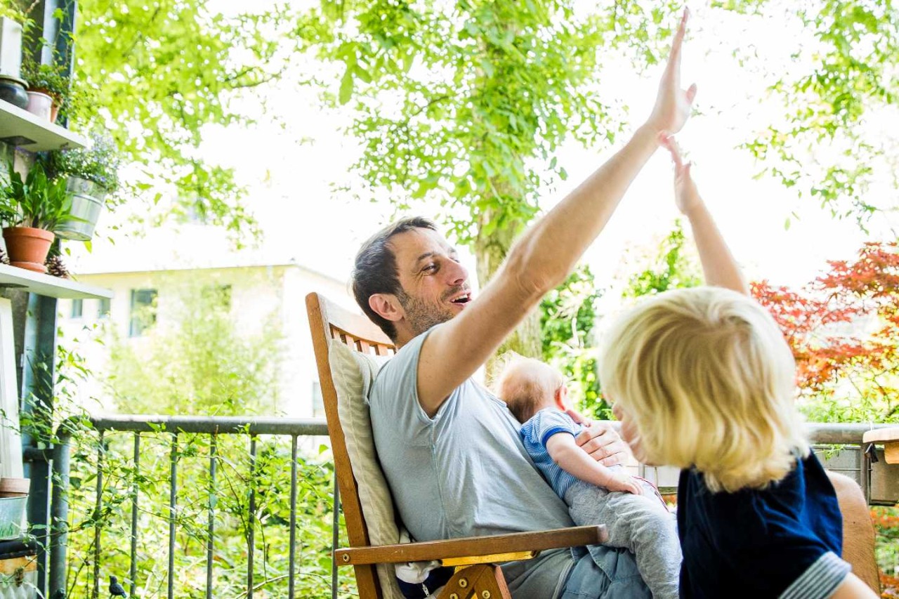 Ein Vater gibt seinem Sohn „High five“ und hält dabei das kleinere Kind auf dem Arm.