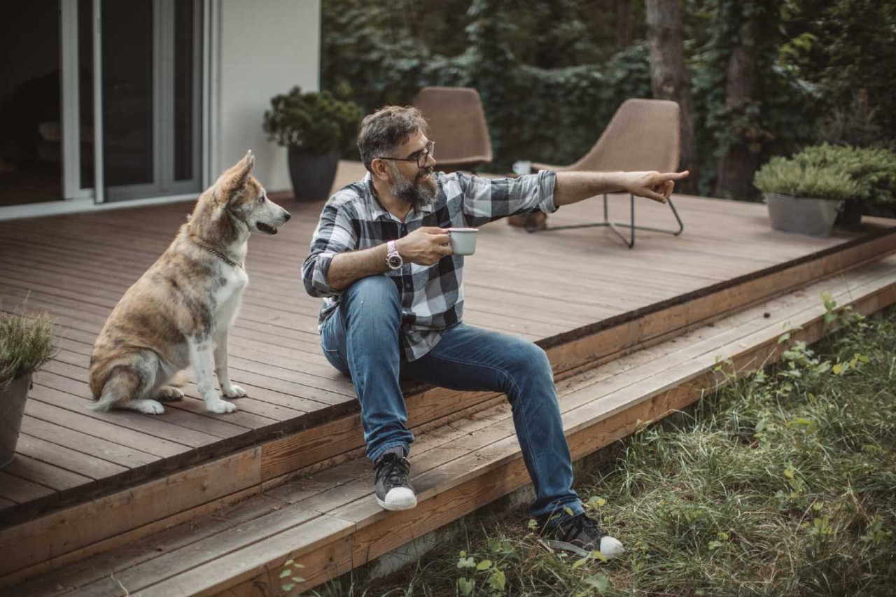 Herrchen sitzt mit Tasse auf Holzterrasse und zeigt Hund etwas in der Ferne.