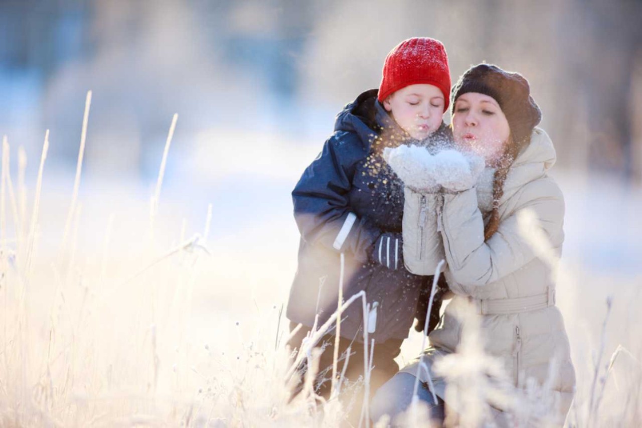 Junge Frau kniet mit einem Mädchen an einem Feldrand und bläst Schnee von den Handschuhen.